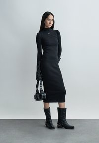 Femme portant une robe midi noire ajustée, des gants noirs longs, des bottes noires montantes ornées de chaînes, tenant un petit sac à main noir, debout sur un sol gris.