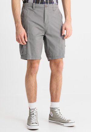 Homme portant un short cargo gris, des chaussettes blanches et des baskets montantes grises, debout devant un fond blanc uni.