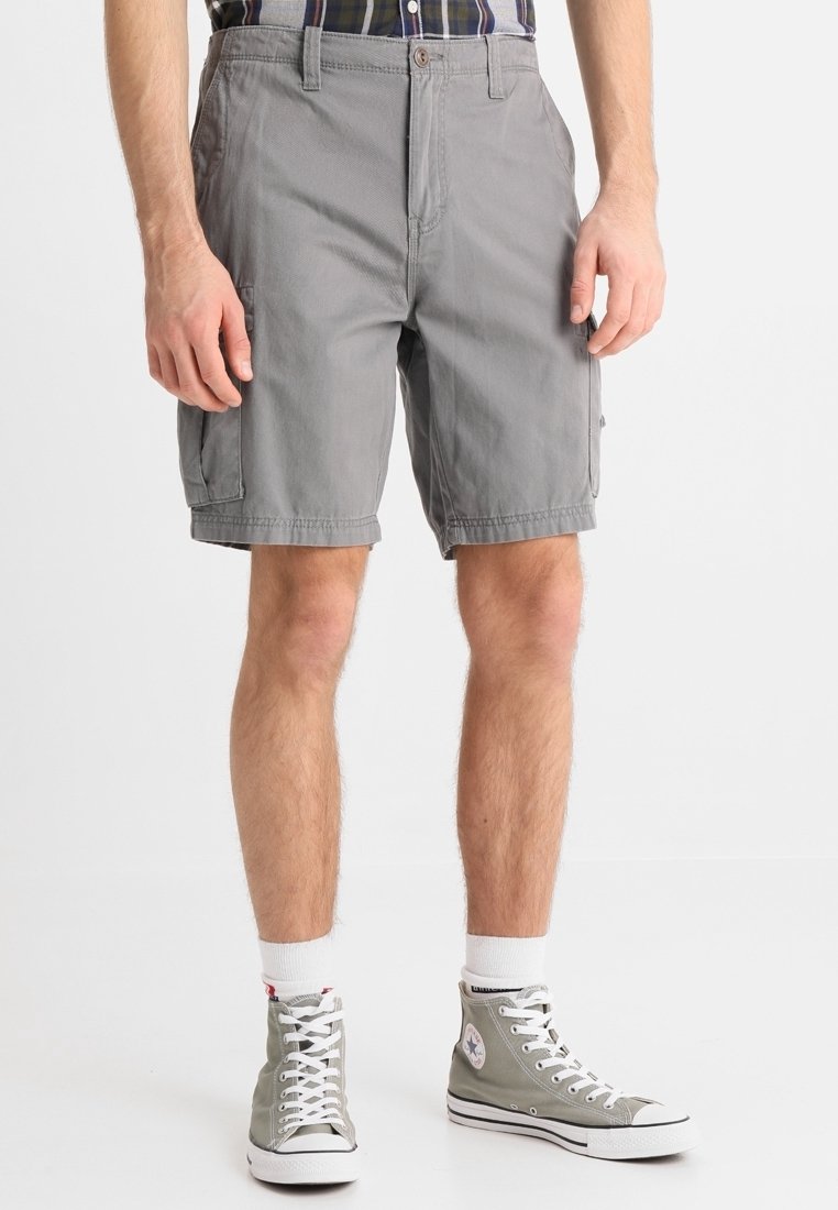 Homme portant un short cargo gris, des chaussettes blanches et des baskets montantes grises, debout devant un fond blanc uni.