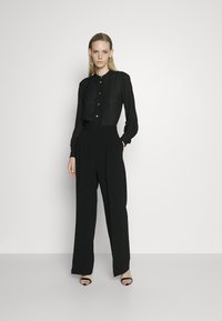 Blouse noire à boutons avec des plis, associée à un pantalon noir taille haute et à jambe large. Accessoirisée avec des boucles d'oreilles circulaires et des escarpins noirs.