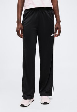 Personne portant un pantalon de survêtement Adidas noir avec des bandes blanches et un logo, assorti à des baskets rose clair et un haut rose clair.