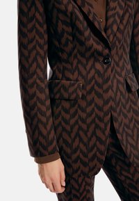 Brown patterned blazer featuring a zigzag design, two front pockets, black button closure, and tailored fit with a smooth texture.