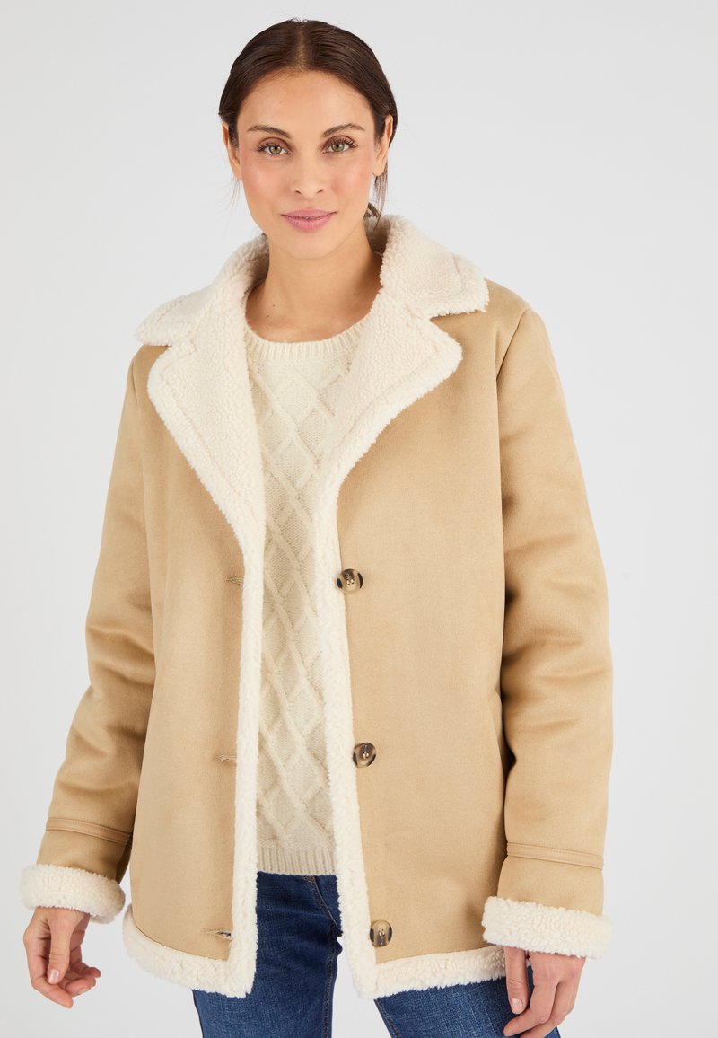 Cappotto beige foderato in shearling con colletto, tre bottoni frontali e polsini in pile, indossato sopra un maglione beige texturizzato e jeans blu.
