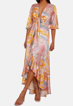 Kobieta ubrana w pastelową sukienkę maxi w abstrakcyjny wzór w kolorach różu, pomarańczu i niebieskiego, z bufiastymi rękawami, wiązaniem z przodu, falbankowym asymetrycznym dołem oraz na nogach cieliste sandały na cienkich paskach.