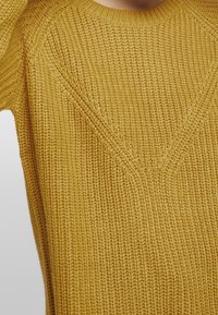 Senfgelbes, gestricktes Sweatshirt mit einem strukturierten, gerippten Muster und Rundhalsausschnitt, das diagonale Linien im Brustbereich aufweist.
