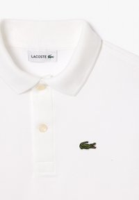 Polo shirt bianco realizzato in tessuto testurizzato, con una placca a due bottoni e un logo di coccodrillo ricamato verde sul lato sinistro del petto.