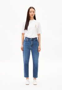 Witte t-shirt met een klein tan logo op de borst, gecombineerd met hoge blauwe denim jeans. Model draagt witte sneakers.