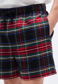 Καρό σορτς με μοτίβο σε μαύρο, κόκκινο, πράσινο και μπλε, με ελαστική μέση και κορδόνι. Το υλικό φαίνεται μαλακό και υφασμάτινο.