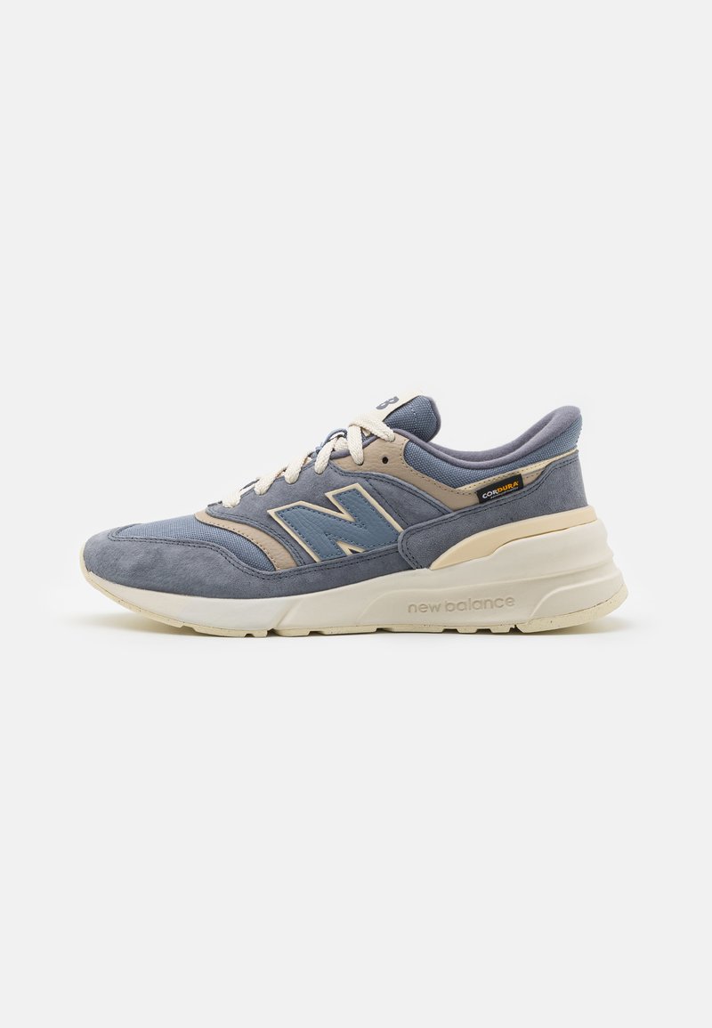 Blaue und beige New Balance Sneaker mit weißen Schnürsenkeln, Obermaterial aus Wildleder und Mesh sowie einer cremefarbenen Sohle mit "new balance"-Schriftzug an der Seite.