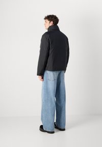 Zwarte gewatteerde jas met hoge kraag, gecombineerd met lichtblauwe wijde jeans. De outfit heeft een strakke, minimalistische uitstraling.