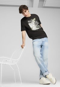 Camiseta gráfica negra con estampado en la pantalla, jeans de mezclilla azul claro y zapatillas blancas. El modelo se inclina contra una silla de alambre blanca.