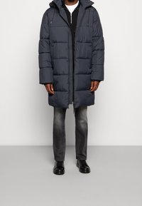Parka grise gonflée avec une fermeture éclair et une capuche, présentant des coutures horizontales. Portée sur un haut noir et associée à un jean gris et des chaussures noires.