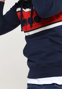 Hoodie bleu marine avec des rayures horizontales rouges et blanches. Dispose d'une poche frontale et de cordons de serrage sur la capuche. Matériau en coton.