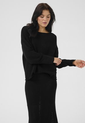 Black knit sweater with a relaxed fit, wide neckline, and long sleeves. Worn with a matching black skirt, showing texture and layered design.