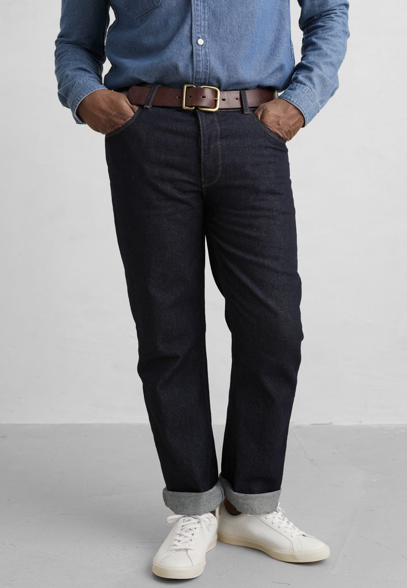 Jeans de mezclilla oscura con un corte recto, dobladillo remangado, costuras amarillas, combinados con una camisa de chambray azul claro y zapatillas blancas. Cinturón marrón con hebilla dorada.