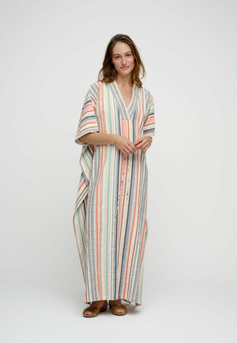 Femme debout, portant une longue robe ample rayée verticalement multicolore avec des manches trois-quarts et des sandales marron, souriant doucement.