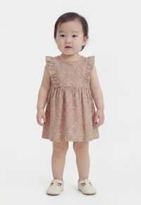 Kleinkind, das ein pinkes, ärmelloses Kleid mit Blumenmuster und Rüschen sowie beige Schuhe trägt und vor einem schlichten weißen Hintergrund steht.