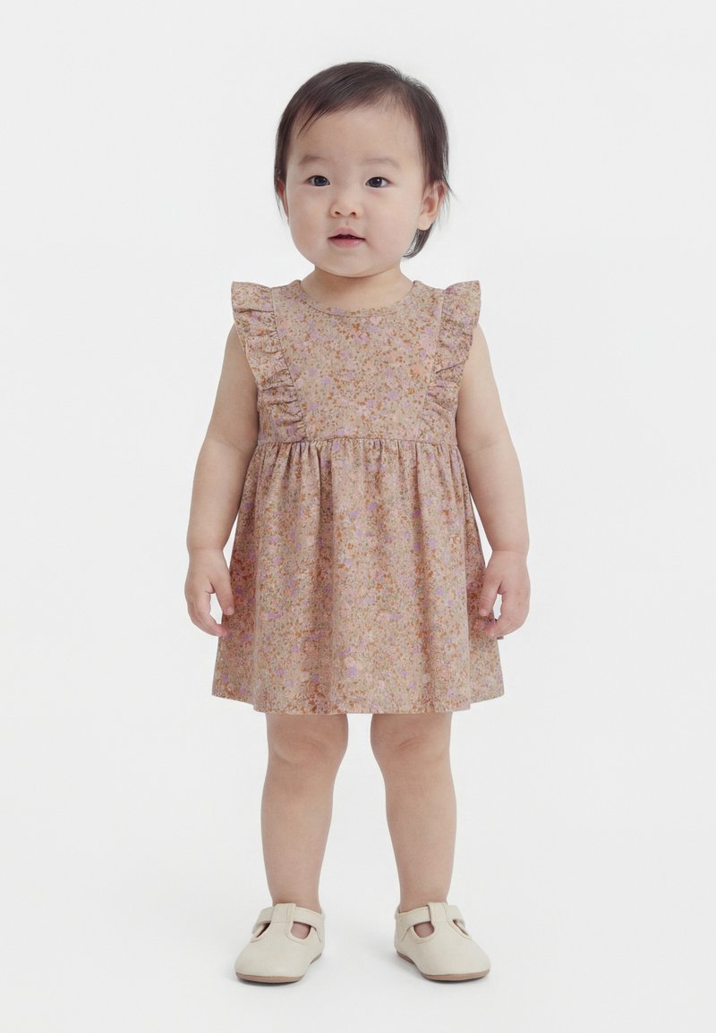 Kleinkind, das ein pinkes, ärmelloses Kleid mit Blumenmuster und Rüschen sowie beige Schuhe trägt und vor einem schlichten weißen Hintergrund steht.