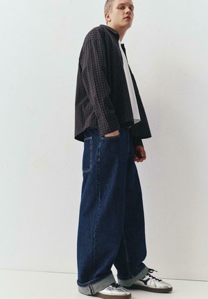 Jeune homme portant une surchemise noire à carreaux, t-shirt blanc, jean bleu large avec revers et baskets argentées, debout devant un mur blanc uni.