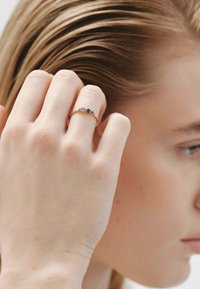 Anillo de oro con tres piedras preciosas alternas: una piedra clara y dos piedras azules, montado en una banda delgada, llevado en una mano suavemente iluminada.