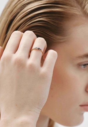 Anillo de oro con tres piedras preciosas alternas: una piedra clara y dos piedras azules, montado en una banda delgada, llevado en una mano suavemente iluminada.