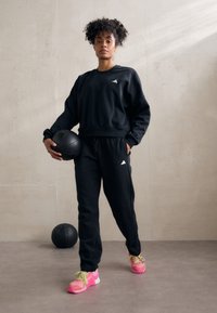 Μαύρη μπλούζα με κουκούλα και φόρμες από μαλακό ύφασμα, με το λογότυπο της Adidas. Φωτεινές ροζ αθλητικές παπούτσια με πράσισα κορδόνια. Κρατάει μια μαύρη μπάλα μπάσκετ.