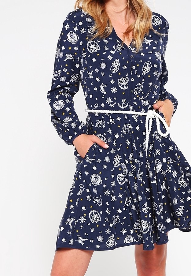 Femme portant une robe bleu marine avec des motifs nautiques et étoiles, manches longues, boutonnée devant et ceinture en corde blanche, mains dans les poches.
