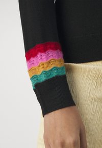 Czarny rękaw z wielokolorowymi prążkami w czerwonym, różowym, musztardowym i turkusowym kolorze, nad ściągaczem, noszony na kremowych, teksturowanych spodniach.