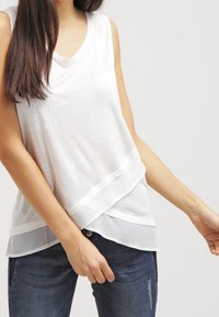Top bianco senza maniche con scollatura a V, design a strati, decorato con motivi circolari ricamati e orlo inferiore trasparente. Combinato con jeans blu.