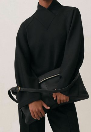 Personne vêtue d'un haut noir à manches longues tenant une grande pochette en cuir noir avec une fermeture éclair et une longue bandoulière à l'avant.
