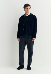 Marineblauwe corduroy jas met voorzakken, gedragen over een donkergroen shirt en grijze broek. Het model staat voor een neutrale achtergrond en draagt zwarte schoenen.