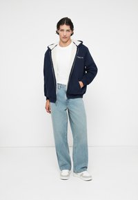 Sweat à capuche zippé bleu marine avec doublure en polaire blanche, poches avant et texte brodé. Associé à un jean bleu clair et des baskets blanches.