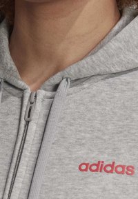 Felpa grigia con zip, realizzata in un tessuto testurizzato. Presenta il logo rosso di adidas, zip grigia e coulisse. Primo piano dell'area del collo.
