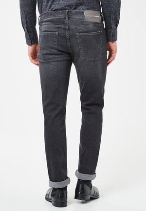 Mężczyzna ubrany w podwinięte ciemnoszare jeansy i czarne eleganckie buty, widziany od tyłu na jednolitym białym tle.