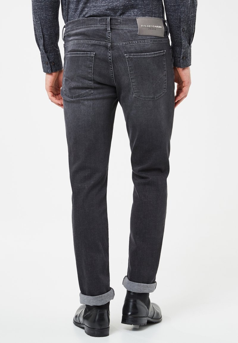 Homme portant un jean gris foncé retroussé et des chaussures de ville noires, vu de dos sur un fond blanc uni.