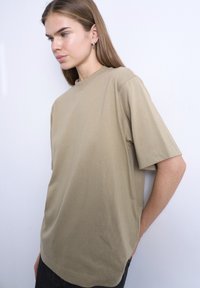 Νεαρό άτομο με μπεζ φαρδιά μπλούζα με κοντά μανίκια, στέκεται μπροστά σε λευκό τοίχο, κοιτάζοντας πλάγια με τα χέρια πίσω από την πλάτη.