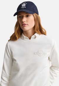 Jeune femme aux longs cheveux portant une casquette bleu marine avec un logo blanc et un sweat-shirt blanc avec un emblème en relief sur la poitrine.