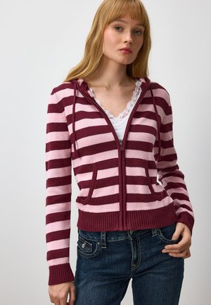 Young woman wearing a maroon and pink striped zip-up hoodie over a white lace-trimmed top and dark blue jeans, standing against a plain background.