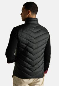 Čierny páperový vest s chevron quiltovaným vzorom, vysokým golierom a zipsovými bočnými vreckami, nosený cez čiernu dlhú tričko s dlhým rukávom.