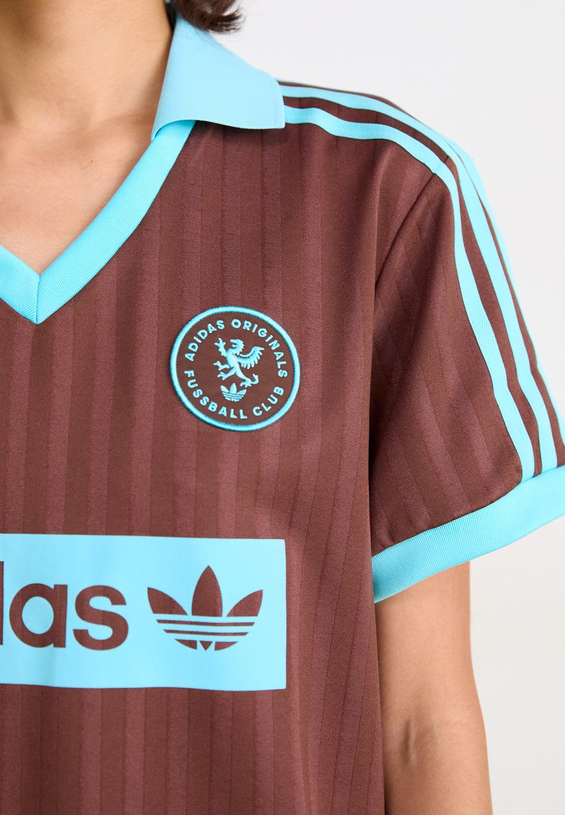 Jersey marrón con rayas verticales. Color azul claro en el cuello y detalles. Presenta un logotipo circular y un gran gráfico de la marca en azul.