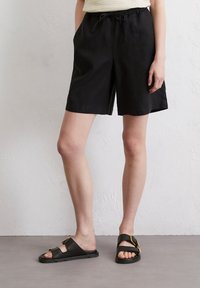 Schwarze Leinen-Shorts mit elastischem Bund und Kordelzug, ausgestattet mit Seitentaschen. Kombiniert mit schwarzen Sandalen mit goldenen Akzenten.