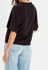 Haut en coton noir à manches courtes, avec un motif de petits points texturé et une bordure en dentelle festonnée le long des manches et de l'encolure.