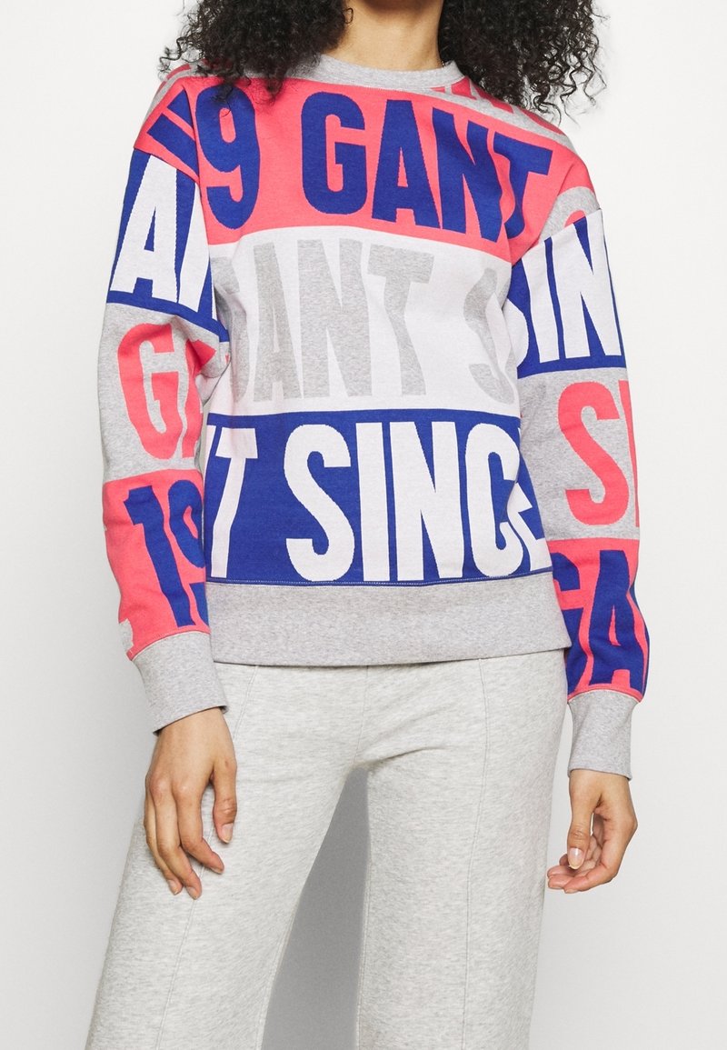Femme portant un sweat-shirt coloré avec des motifs de texte en rouge, bleu et blanc vif, associé à un pantalon gris clair sur un fond uni.