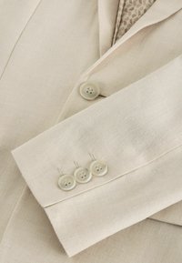 Veste de costume beige avec quatre boutons assortis sur la manche et un bouton près du revers, présentant un tissu texturé et une doublure à motifs.