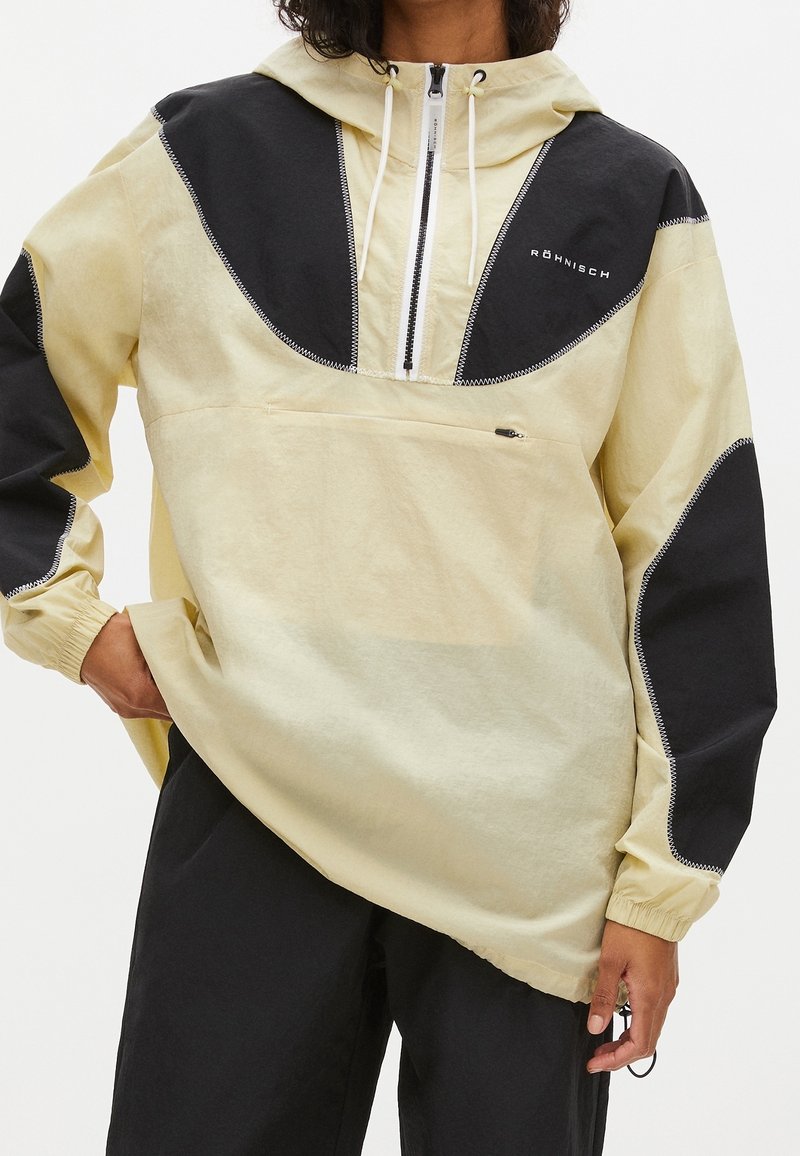 Personne portant un sweat à capuche beige et noir avec une fermeture éclair à l'avant et une poche zippée, associée à un pantalon noir.