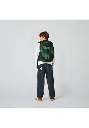 Junge mit einem grünen und schwarzen gemusterten Rucksack, weißem Langarmshirt, dunkler weiter Hose mit einem Smiley-Patch und braunen Schuhen, steht mit dem Rücken zur Kamera.