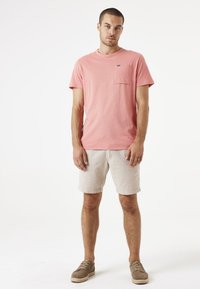 Camiseta de algodón rosa con un bolsillo en el pecho, combinada con pantalones cortos de textura beige claro. El modelo lleva zapatos marrones con cordones. Diseño simple y casual.