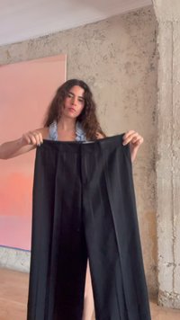 Femme aux longs cheveux bouclés tenant un pantalon noir à jambes larges dans une pièce aux murs beiges texturés et une peinture abstraite rose.