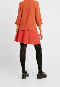 Roter ausgestellter Rock mit horizontalen Nähten, kombiniert mit einer orangefarbenen Bluse mit 3/4-Ärmeln, schwarzen Strumpfhosen und schwarzen Schuhen mit dickem Absatz.