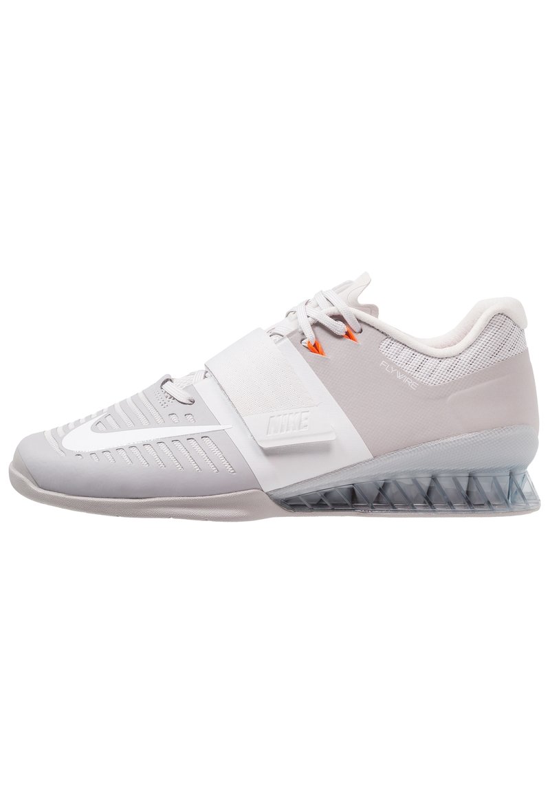 Grå træningssko med en mesh-overdel, en segmenteret gennemsigtig sål og en rem over midtfoden. Har orange detaljer og teksturerede sektioner.